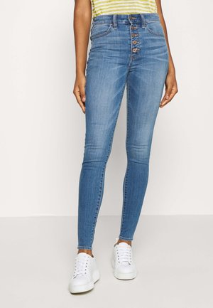 Vrouw draagt lichtblauwe hoge taille skinny jeans met vijf zichtbare knopen en witte platformsneakers, staand tegen een effen achtergrond.