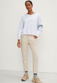 Witte cropped sweatshirt met een V-hals en blauwe letters op de mouw. Gecombineerd met lichtbeige broek en tan kleurige sportschoenen.