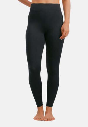 Kvinde iført sort højtaljede leggings, stående barfodet med den ene hånd hvilende nær hoften mod en ensfarvet baggrund.