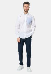 Camicetta bianca con taschino blu a fantasia, abbinata a pantaloni navy e sneakers bianche. Tessuto liscio, vestibilità moderna.