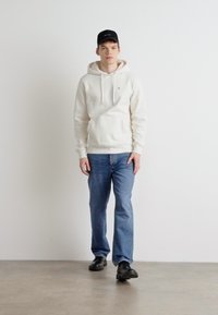 Sudadera crema con cordones, jeans de color azul claro y zapatos negros. La sudadera presenta un pequeño logo en el pecho y una textura suave.