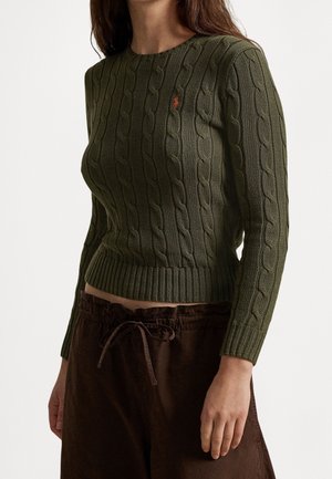 Femme portant un pull en maille torsadée vert olive ajusté et un pantalon ample marron foncé avec une taille à cordon.