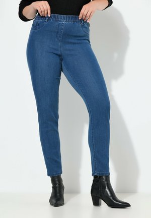 Jeans de mezclilla azul con cintura alta elástica, diseño ajustado y largo hasta el tobillo, combinados con botas de tacón negras hasta el tobillo.