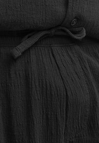 Lähikuva mustasta teksturoidusta kankaasta, jossa on solmittu vyö ja pieni musta nappi vaatteen vyötäröllä.