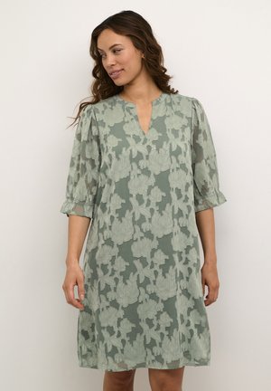Sage green dress with a V-neck and sheer, patterned overlay featuring floral embroidery. Three-quarter puff sleeves with ruffled cuffs.