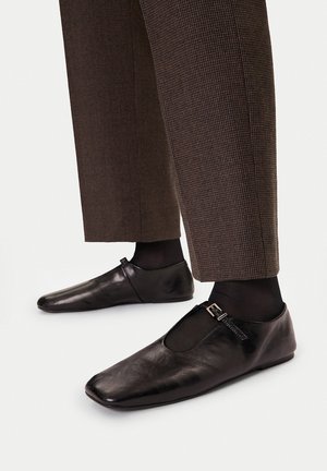 Černé kožené baleríny s T-ramínkem a přezkou, nošené s černými punčocháči a hnědými kostkovanými zkrácenými kalhotami na bílém pozadí.