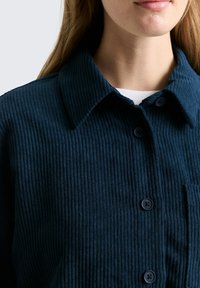 Navyblauwe ribfluwelen shirt met een kraag, knoopsluiting aan de voorkant en een zak. De stof heeft een verticale ribstructuur en een zachte afwerking.