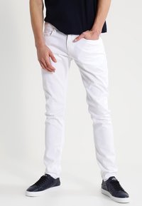 White denim jeans with a slim fit and five-pocket design, worn with navy sneakers. The fabric has a smooth texture and slight stretch.