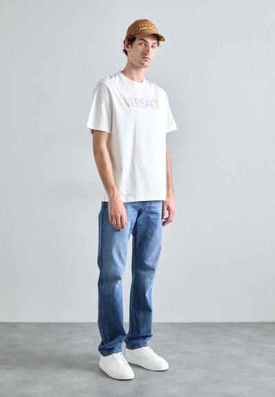 Tricou alb din bumbac cu logo-ul roz "VERSACE", asortat cu blugi albaștri și adidași albi. Modelul poartă o șapcă maro cu logo.