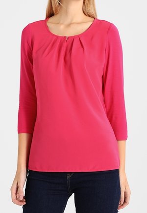 Femme portant un haut à manches longues rose vif avec un col rond et de légers plis près du col, associé à un jean bleu foncé.
