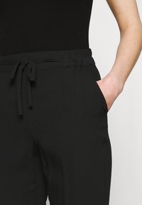 Nära bild av en person som bär svarta byxor med elastiskt midjeband och dragsko, med handen placerad i den högra fickan, mot en enfärgad bakgrund.