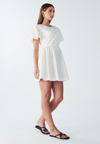 Biele krátke šaty s krátkymi rukávmi a priliehavým korzetom, s rozšírenou sukňou. Hladká textúra, minimalistický dizajn, doplnené čiernymi plochými sandálmi.