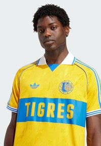 Gul sportstrøje med blå vandret bånd og "TIGRES" tekst. Lavet af glat stof, med tekstureret mønster og Adidas-logo på brystet.