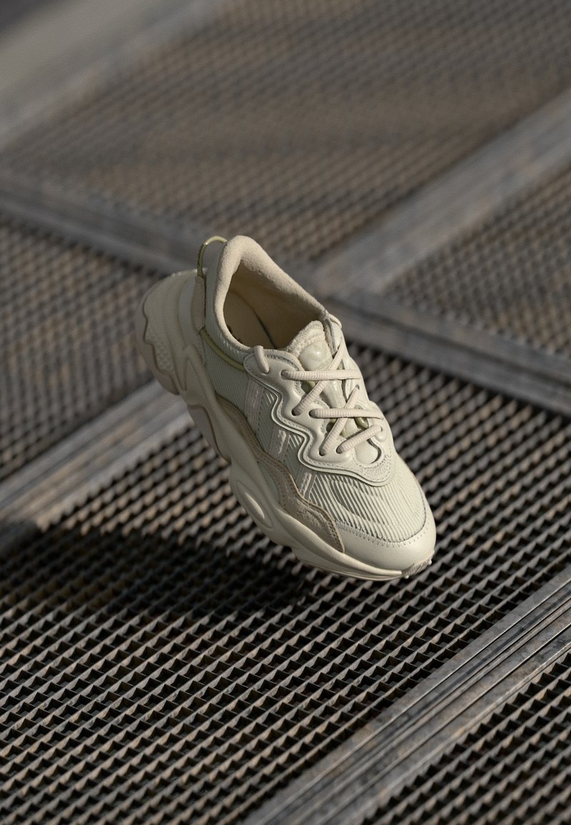 Beige chunky sneakers med texturerad tyg och tjock sula placerade på en metallgaller yta med skuggad belysning.