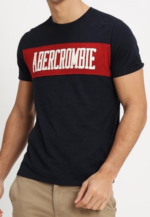Hombre con una camiseta negra con una franja horizontal roja y texto blanco "ABERCROMBIE" en el pecho, combinado con pantalones beige.