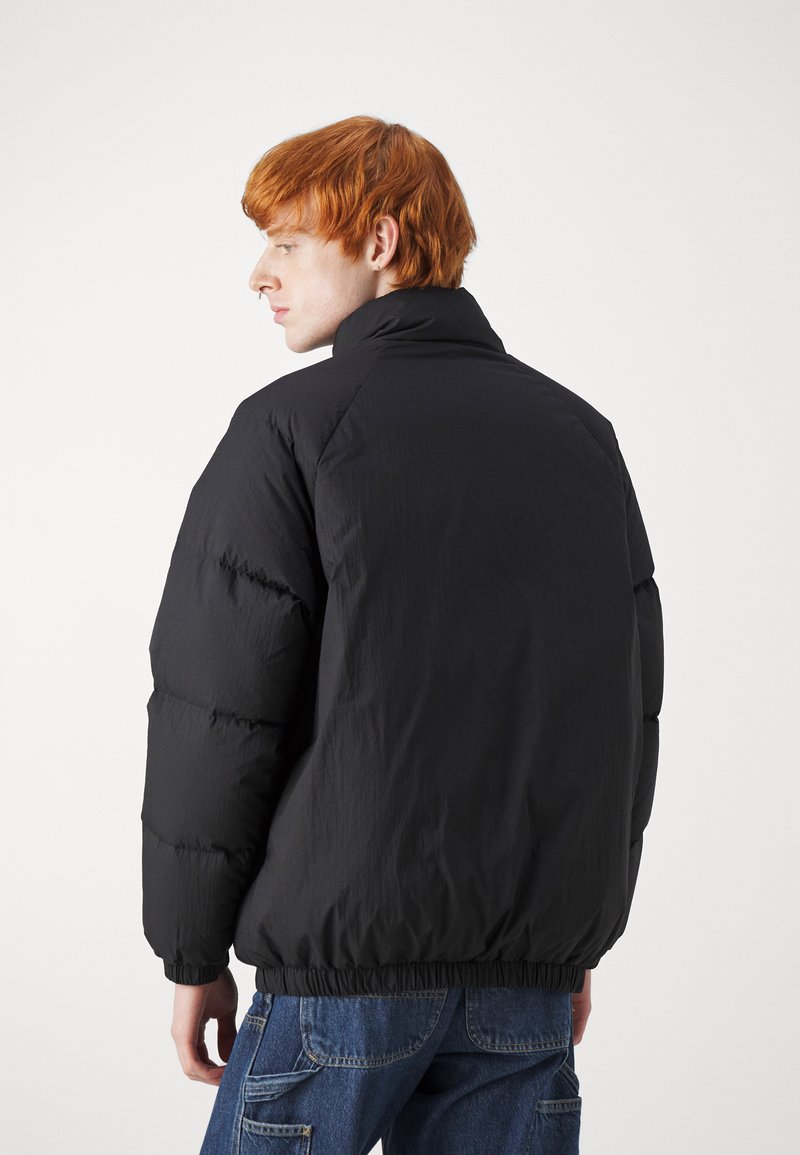 Schwarze Pufferjacke mit hohem Kragen, mit strukturierter Oberfläche und elastischem Saum, getragen über blauer Jeans.
