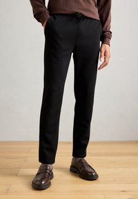 Hombre que lleva pantalones de algodón negros con cordón, mocasines marrones y una camiseta de manga larga marrón, de pie sobre un suelo de madera clara contra una pared neutral.