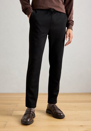 Hombre que lleva pantalones de algodón negros con cordón, mocasines marrones y una camiseta de manga larga marrón, de pie sobre un suelo de madera clara contra una pared neutral.