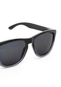 Mustat muoviset aurinkolasit, joissa on tummat linssit ja paksu, kulmikas kehysmuotoilu, sileillä pinnoilla ja hieman kaartuvilla sankoilla.