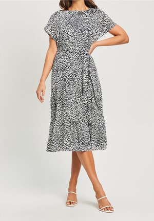 Kobieta ubrana w czarno-białą midi sukienkę w cętki z krótkimi rękawami i paskiem w talii, w zestawieniu z białymi sandałkami na obcasie z paseczkami.