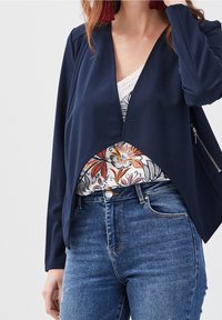 Femme portant une veste bleu marine ouverte sur un haut fleurie à bordure en dentelle, associée à un jean taille haute bleu et des boucles d'oreilles à pompons rouges.