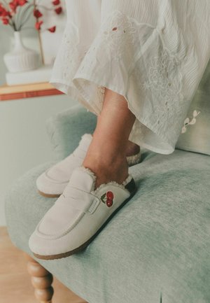 Feet wearing white slip-on slippers with fur lining and cherry embroidery, resting on a light green cushioned chair. White embroidered dress hem visible.