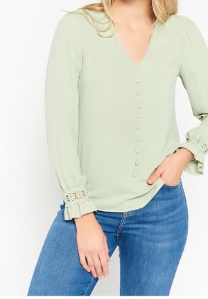 Femme portant un chemisier vert clair à col en V avec des boutons cachés et un détail en dentelle au poignet, associé à un jean bleu, main dans la poche.