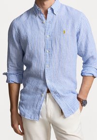 Světle modrá a bílá pruhovaná košile na knoflíky s dlouhými rukávy a límcem, s malým žlutým logem. Vyrobena z prodyšného materiálu.