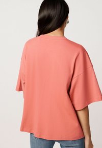 Koraalkleurig oversized T-shirt met korte mouwen, ronde halslijn en een gladde textuur. De zoomlijn ligt op heuphoogte; geen zichtbare patronen of accenten.