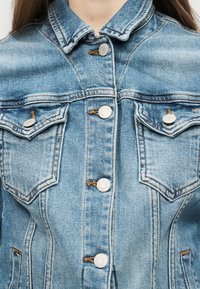 Κοντινό πλάνο ενός ατόμου που φοράει ανοιχτό μπλε τζιν μπουφάν με μεταλλικά κουμπιά και δύο τσεπάκια στο στήθος με κουμπί.