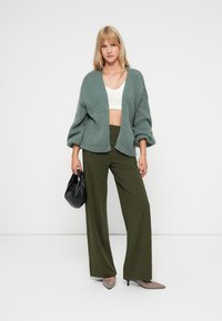 Cardigan en tricot vert à coupe décontractée et manches larges, associé à un pantalon large olive et un sac à main noir. Haut court neutre en dessous.