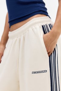 Nahaufnahme einer Person in cremefarbenen Sweatpants mit marineblauen Streifen und Logo, Hand in der Tasche, kombiniert mit einem marineblauen Cropped-Top.