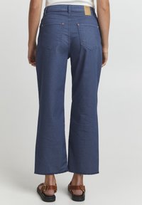 Jeans de denim de pierna ancha en azul oscuro, con un dobladillo sin acabar, bolsillos traseros y un parche de cuero marrón en la cintura. Textura suave.