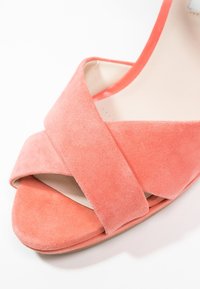 Sandale en suède corail avec un design à bandes croisées, bout ouvert et doublure intérieure lisse. La semelle est plate avec une forme arrondie.