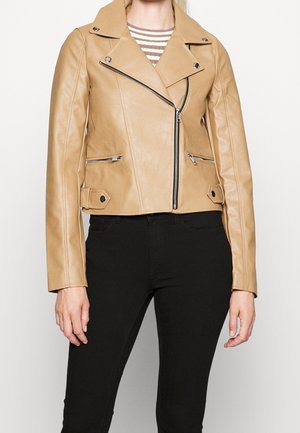 Vrouw draagt een kort lichtbruine leren jack met zilveren ritsen over een gestreept shirt en nauwsluitende zwarte broek.