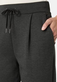 Černé pletené jogger kalhoty mají stahovací pás, záhyby a boční kapsy, a vyznačují se měkkým strukturovaným materiálem.