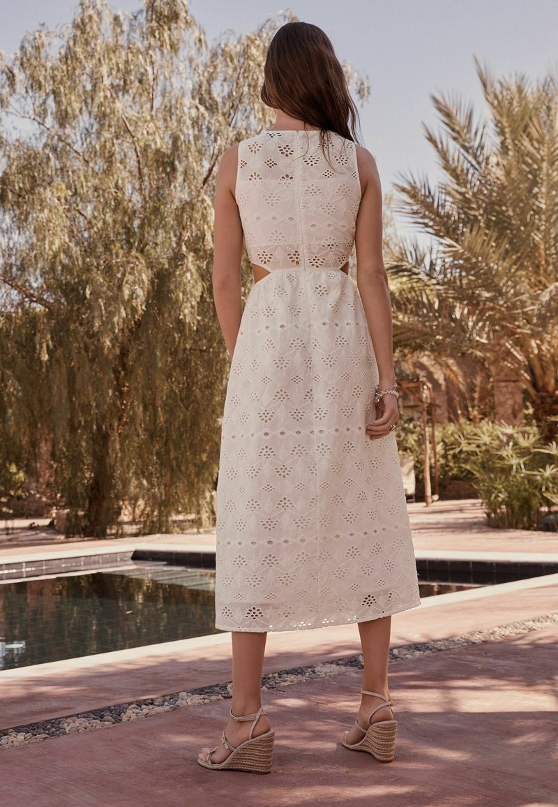Femme aux longs cheveux bruns portant une robe midi blanche sans manches à œillets et des sandales compensées beiges, debout près d'une piscine en plein air avec des arbres en arrière-plan.