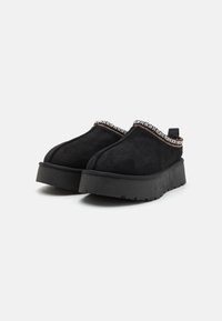 Scarpe slip-on nere in suede con suole in piattaforma nera texturizzata. Presentano un bordo decorativo intrecciato attorno al colletto e una linguetta sul tallone.