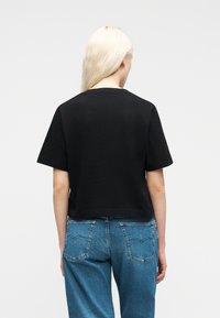 Ξανθός/ή φοράει μαύρο κοντομάνικο πουκάμισο και μπλε τζιν, στέκεται με την πλάτη γυρισμένη προς ένα απλό λευκό φόντο.