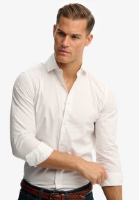 Witte knoopsluiting shirt met lange mouwen, opgerold bij de manchetten. Gemaakt van zachte stof, met een subtiele textuur en een klein logo op de borst.