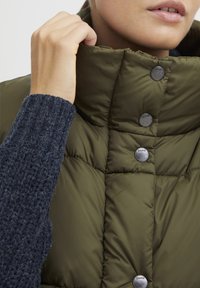 Vest verde măsliniu căptușit cu guler înalt, prins cu butoane metalice argintii. Pulover tricotat texturat de culoare albastru închis vizibil dedesubt.