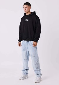 Zwarte hoodie met een witte logo, gecombineerd met lichtblauwe distressed jeans met sterpatronen en grijze sneakers. Casual fit.