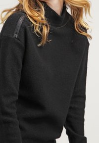 Pullover nero a maglia con collo alto. Presenta una texture liscia, maniche aderenti e due dettagli neri lucidi sulle spalle.