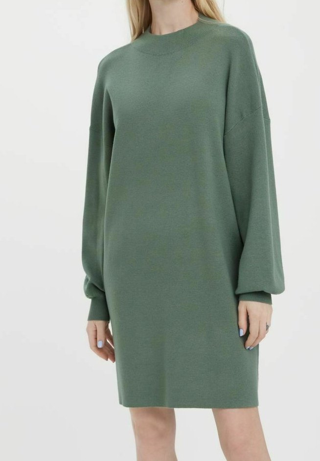 Femme portant une robe tricotée ample à manches longues de couleur vert sauge avec des poignets côtelés et un col rond haut, sur un fond blanc uni.