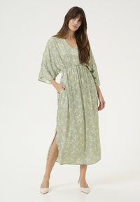 Dolga zelena obleka s cvetličnim vzorcem, z V-izrezom, zategnjenim pasom in stranskimi razporki. Lahka tkanina, sproščen kroj in kratki rokavi.