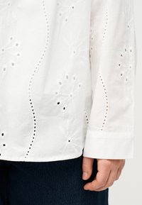 Άσπρο πουκάμισο με μακριά μανίκια και κεντημένη λουλουδάτη διακόσμηση, φορεμένο πάνω από σκούρο παντελόνι, με εμφανές το μανίκι και το χαλαρό χέρι στο πλάι.