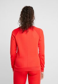 Červená sportovní mikina s ribbed texturou, kulatým výstřihem a přiléhavým střihem. Obsahuje kontrastní texturované ramenní panely.
