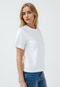 Witte t-shirt met korte mouwen en een ronde hals, met verfijnde sprankelende accenten aan de voorkant. Gecombineerd met blauwe spijkerbroek.