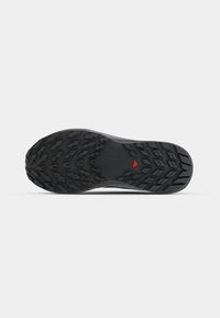 Semelle noire d'une chaussure avec un motif de crampons détaillé et un accent triangulaire rouge près du centre. Fabriquée en caoutchouc durable.