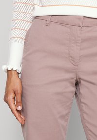 Ljust rosa chinos med en slät finish, med en framficka, bältesömmar och en slim fit-design. Texturerad vit ärm delvis synlig.